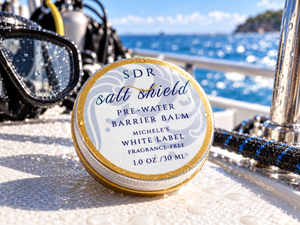 Round tin of SDR Salt Shield Pre-Water Barrier Balm, Michele’s White Label Fragrance-Free, on a wet dive boat deck beside scuba gear with the ocean in the background.